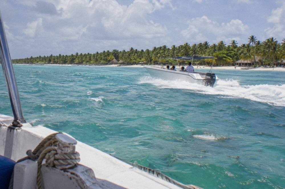 A white speedboat cutting across calm turquoise water toward a palm-lined island shoreline
